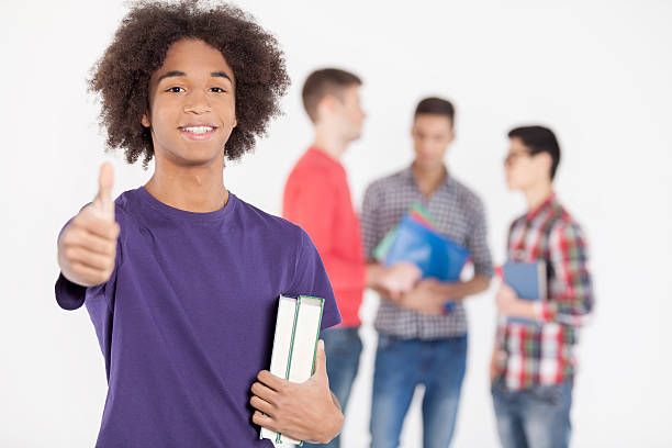 Cheerful African teenage boy holding books and gesturing while his friends standing on background