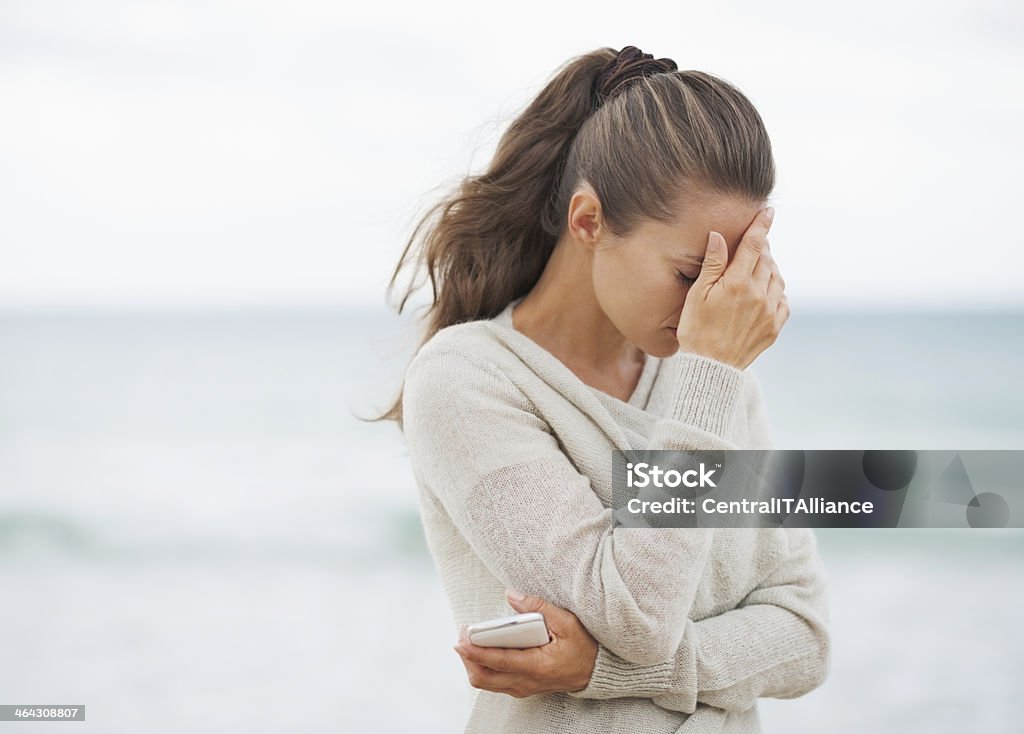 stressed young woman in sweater on beach with cell phone Stressed young woman in sweater on beach with cell phone Sadness Stock Photo