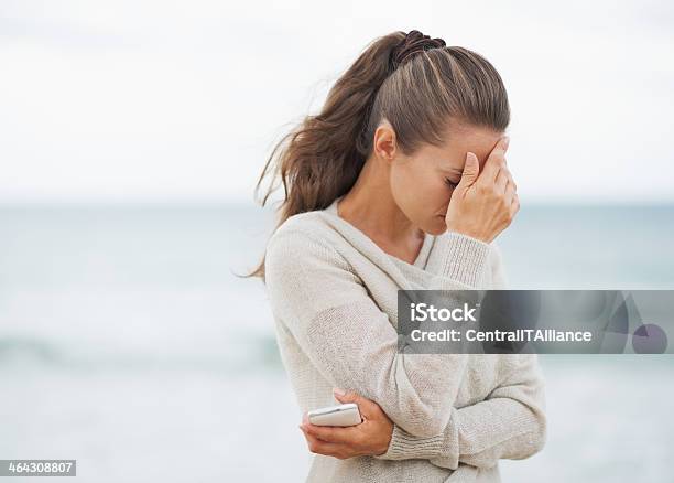 Stressed Young Woman In Sweater On Beach With Cell Phone Stock Photo - Download Image Now