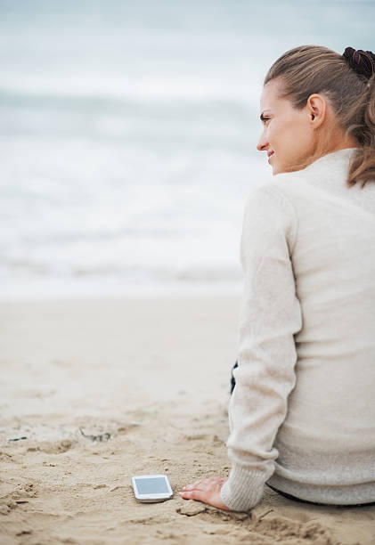 istock Young woman with cell phone sitting on lonely beach 464308861