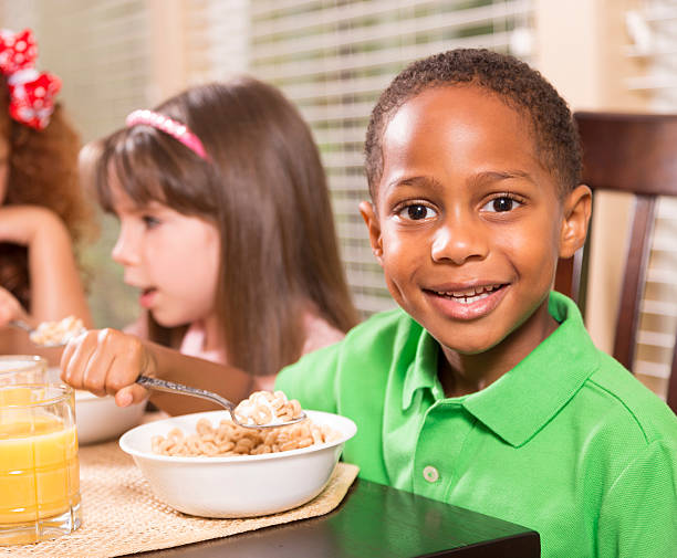 Food:  Three multi-ethnic children enjoying breakfast together. Three multi-ethnic friends have cereal and orange juice for breakfast.  African descent boy in foreground.  School or home setting.  boys bowl haircut stock pictures, royalty-free photos & images
