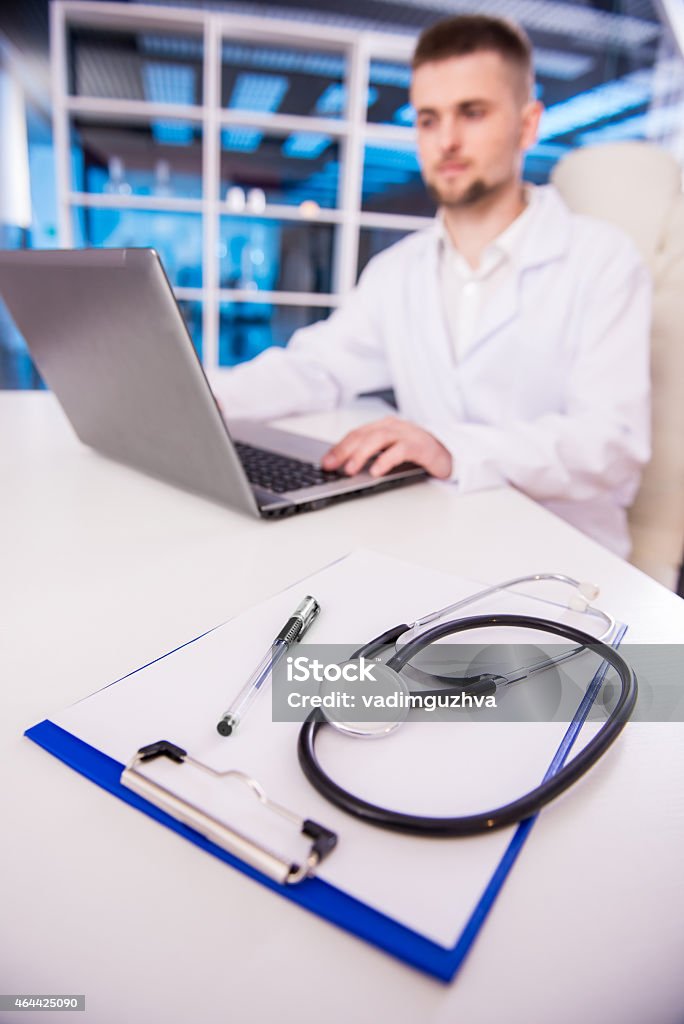 Doctor Young doctor is working in his office. Focus on stethoscope on the table.Portrait of friendly male doctor is looking at the camera and smiling. 2015 Stock Photo