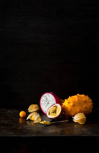 Four types of exotic tropical fruit placed together on a very dark background for a still life effect with copy space on top. The fruits displayed are physalis (or Cape gooseberry), pitaya (or dragonfruit), passionfruit and melano (or horned melon or kiwano).