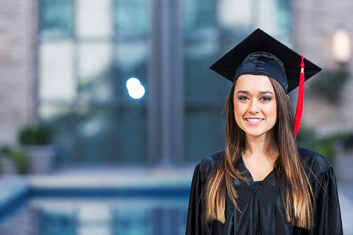 Portrait of a young woman wearing a black graduation cap and gown, standing outdoors in front of the school building.  She is mixed race, Hispanic and Caucasian, with long, straight, dark brown hair.