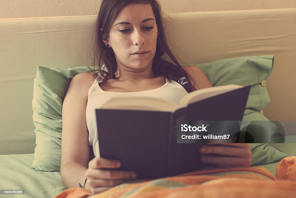 Young Woman Reading a Book on the Bed Young Woman Reading a Book on the Bed. She is Alone, wearing a white Undershirt. The bed has green and orange Sheets. The Girl is Holding the Book with both Hands while Lying down on the Bed. 2015 Stock Photo