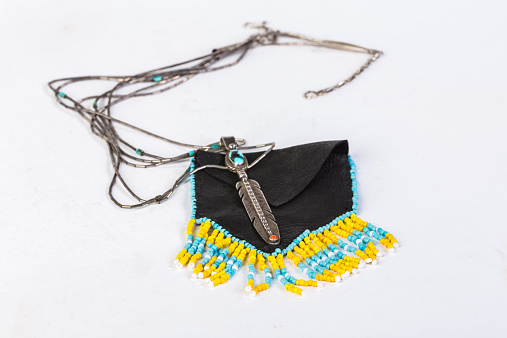 Native American beaded medicine bag and silver feather pendant with turquoise and coral bead against plain background.