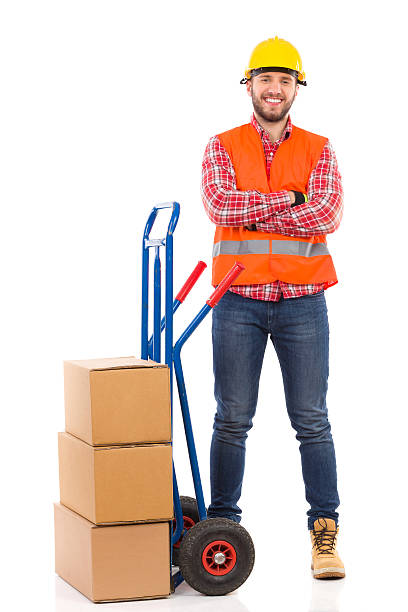 Smiling mover with a push cart. Smiling man in yellow hardhat and orange reflective vest posing with arms crossed next to delivery cart. Full length studio shot isolated on white. mover delivery person box hand truck stock pictures, royalty-free photos & images