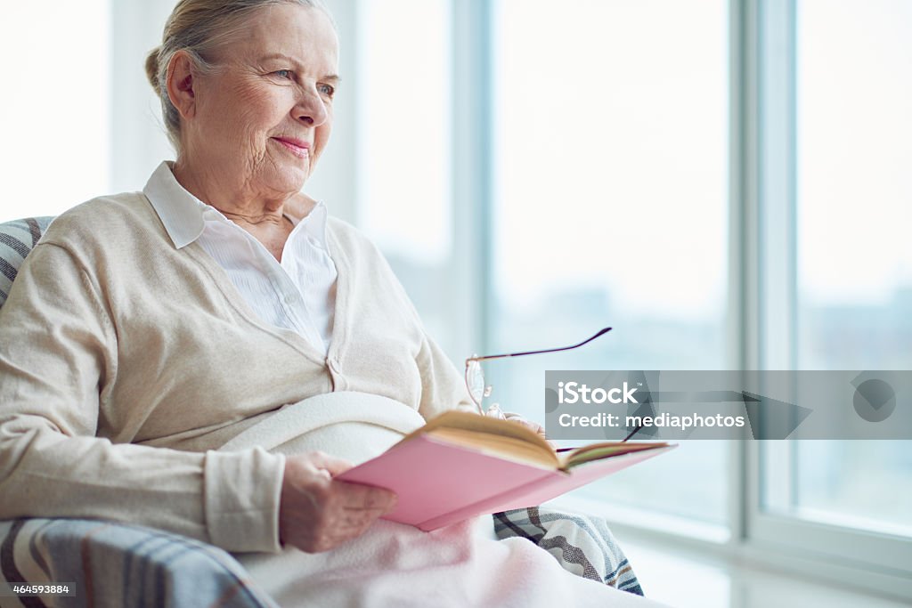 Reading and dreaming Smiling senior woman holding open book and looking through window 2015 Stock Photo