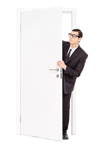 Full length portrait of a businessman peeking through an opened door isolated on white background