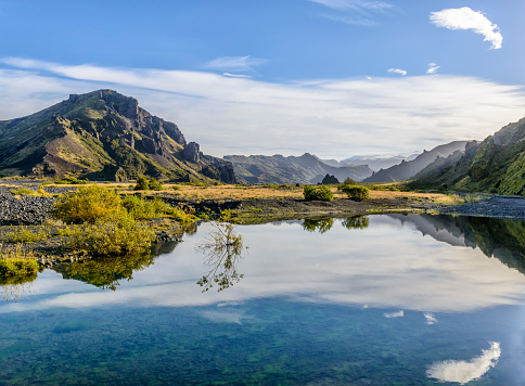 A landscape photograph of the Krossa river in the Thorsmork region in Iceland. There is a mountain range which is reflected in the river. The water is very still and clear. It is quite cloudy but the blue sky is visible. There are clumps of green grass.