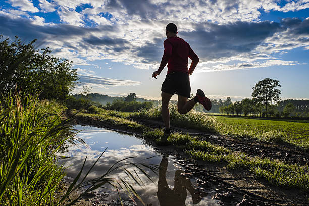 A young man doing trail running in the countryside and jumping over the water