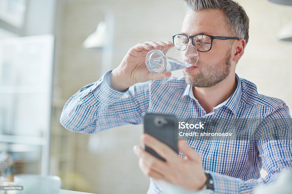 Refreshment during work Businessman in casualwear drinking water while reading sms or dialing number on cellphone Water Stock Photo