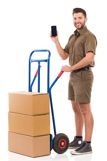 Courier showing a smart phone Smiling messenger standing with a push cart and presenting a cell phone. Full length studio shot isolated on white. mover delivery person box hand truck stock pictures, royalty-free photos & images