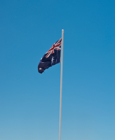 Flying Australian flag on a long white flagpole. Sky is gradient blue with no clouds. Some of the stars on flag can be partially seen.