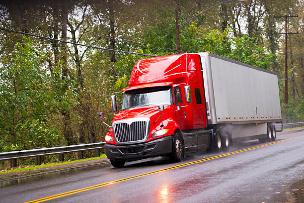 Modern red glossy in rain semi truck trailer raining road Big red semi truck shiny and wet from the rain with the reflection of light with a long distance measuring trailer with dust rain under the wheels and reflection of headlights on a wet road passing by green trees. truck rain stock pictures, royalty-free photos & images