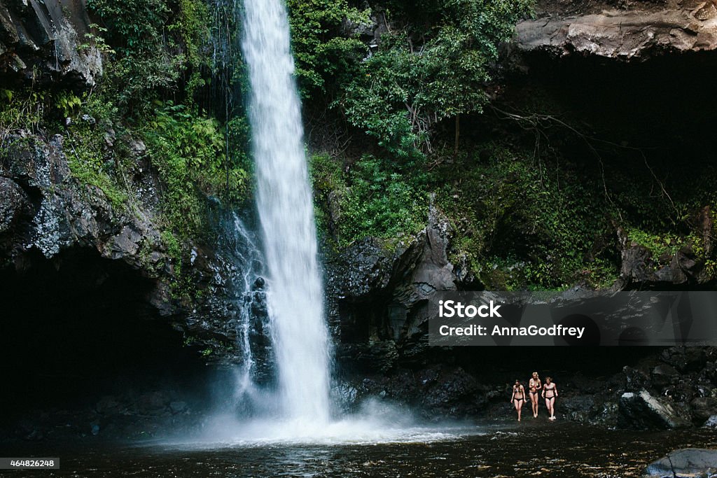 Three Women by a Waterfall in Hawaii Three women enter the dark water of a pool created by a large waterfall. Photographed in Hana, Hawaii.  Waterfall Stock Photo
