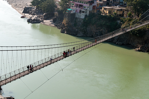 Rishikesh, India - December 9, 2014:  Laxman Jhula bridge over Ganges river.  People crossing footbridge. Rishikesh is  World Capital of Yoga,  has numerous yoga centres that also attract tourists