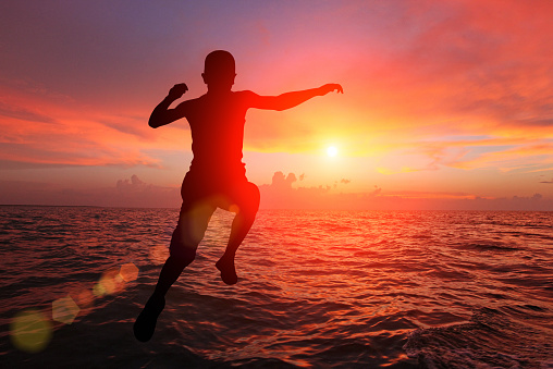 Silhouette of young boy with arm raised jumping into sea water at sunset, while sun shines behind him creating lens flare: courage and determination. Unrecognizable person. Dramatic sky with cloudscape.