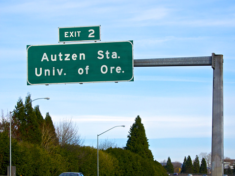 A Freeway Exit sign showing directions to Autzen Stadium and the University of Oregon.  This is a winter, Blue Sky day in Oregon. A few trees are visible.