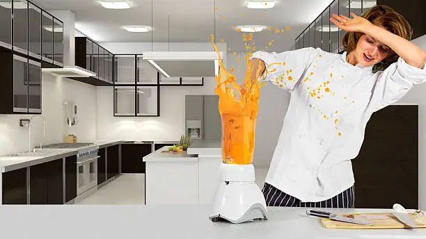Photo of a young female chef making an mess with a blender.  The pretty caucasian female is wearing a white chef uniform. She is in a commercial or domestic kitchen.  The blender is spilling juice because she forgot to put the cover lid on.  The image depicts failure at the workplace in the food and service industry.  