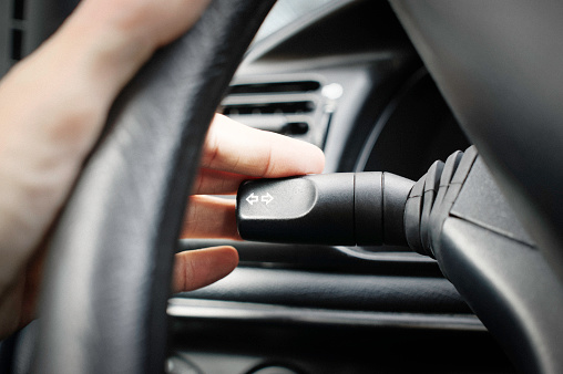 Close-up of a left hand setting the left blinker on the car by pushing down the switch. Some vignetting and digital grain added