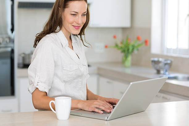 istock Smiling young woman using laptop in kitchen 464861616