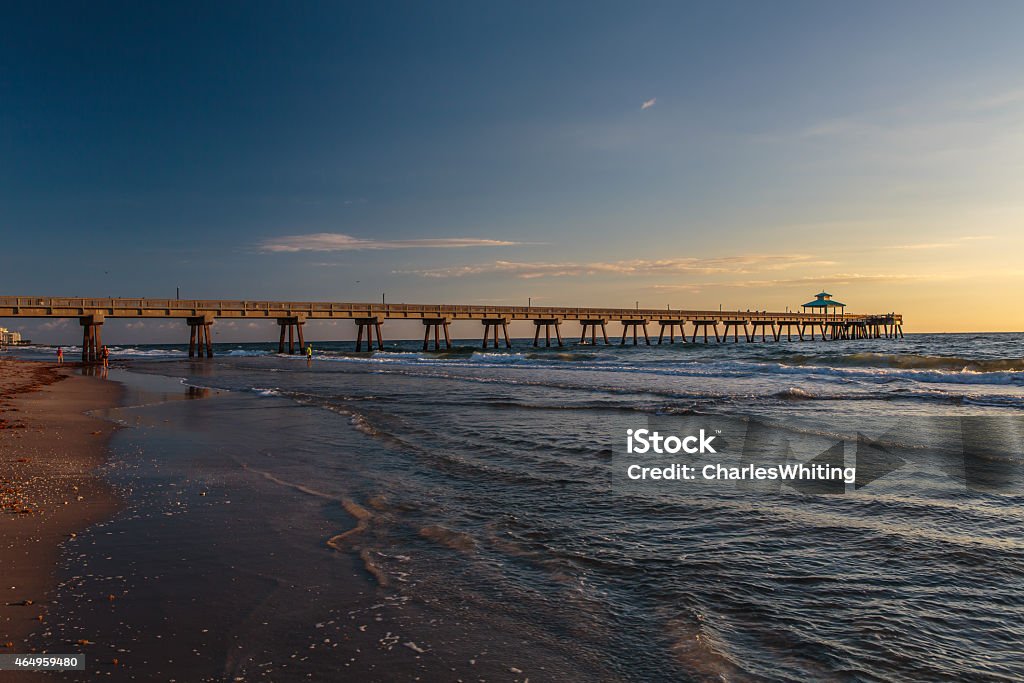 Fishing Pier at Sunrise with Blue Sky and White Clouds The first light of day shines on a fishing pier in Deerfield Beach Florida. The blue sky, white clouds, and ocean reflect the rich warm light of the morning. 2015 Stock Photo