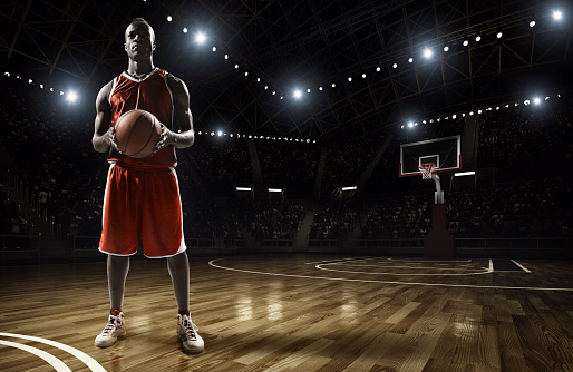 Basketball player standing holding basketball on an indoor floodlit basketball court full of spectators.