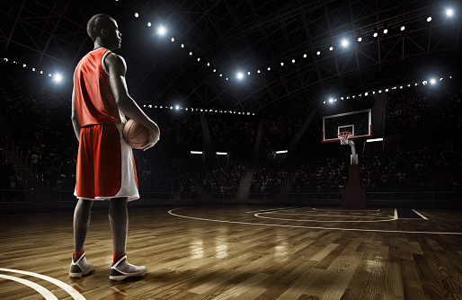 Basketball player standing holding basketball on an indoor floodlit basketball court full of spectators.