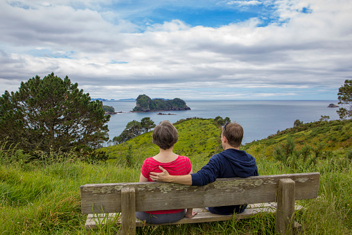 A men and a women sitting on a beach overlooking a beautiful ocean view. Taking in a gorgeous scene near Cathedral Cove, New Zealand