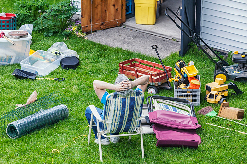 A mature, senior adult woman doing spring cleaning is a bit frazzled with frustration at all the remaining work. She is running her hands through her hair as she rests in a folding chair after completely emptying the back yard shed of all sorts of outdoor household items, toys and gardening equipment.