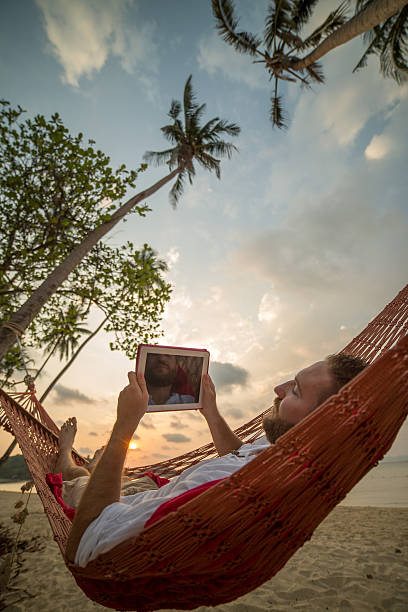 Man uses wireless technology on hammock at sunset Young man lying down on hammock relaxing and using a digital tablet. Sunset on Koh Mak Island, Thailand. hammock men lying down digital tablet stock pictures, royalty-free photos & images