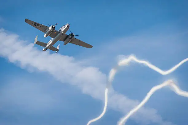 A vintage WWII North American B-25 "Mitchell" bomber flies through a web of contrails left by fighter planes on a low-level pass