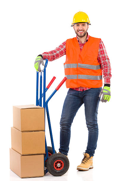 Mover posing Smiling man in yellow hardhat and orange reflective vest posing with a delivery cart and looking at camera. Full length studio shot isolated on white. mover delivery person box hand truck stock pictures, royalty-free photos & images