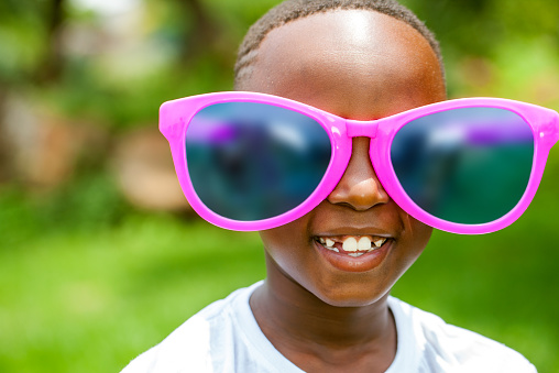 Close up face shot portrait of cute African boy wearing huge over sized sun glasses outdoors.