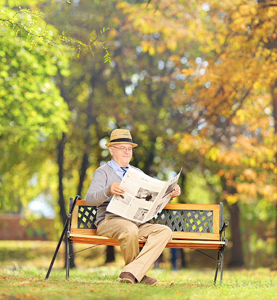 istock Senior man seated on a bench reading newspaper in park 465135461