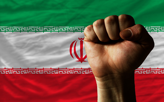 complete national flag of iran covers whole frame, waved, crunched and very natural looking. In front plan is clenched fist symbolizing determination