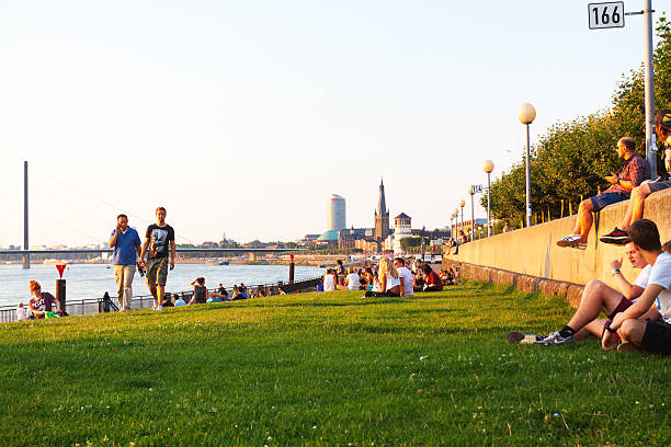 Düsseldorf, Germany - July 23, 2014: Summer evenign and sunset at river Rhine in Düsseldorf. Many people are relaxing on promenade and on grass. In background is center of Altstadt with church and lighthouse.
