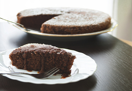 A slice of French style chocolate cake is seen in the foreground sitting on a white plate. In the background, out of focus, is the rest of the cake on a plate. Both plates visible in natural light and are on a dark brown, wooden table. Delicious chocolate cake.