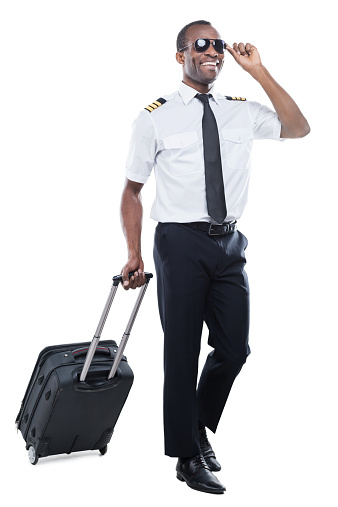 Happy African pilot in uniform walking and carrying suitcase while being isolated on white background