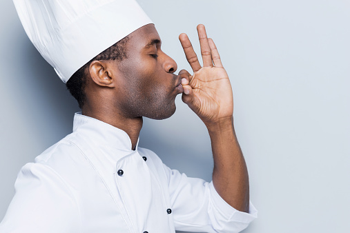 Side view of confident young African chef in white uniform keeping eyes closed and gesturing while standing against grey background