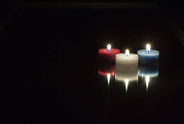 Three candles arranged on a mirror with their flames lit. One red, one blue and one white. The mirror is black as well as the background. Each candle and flame are reflected back in the mirror so the appearance is six flames and candles. The orientation is the middle right leaving plenty of room for copy. 