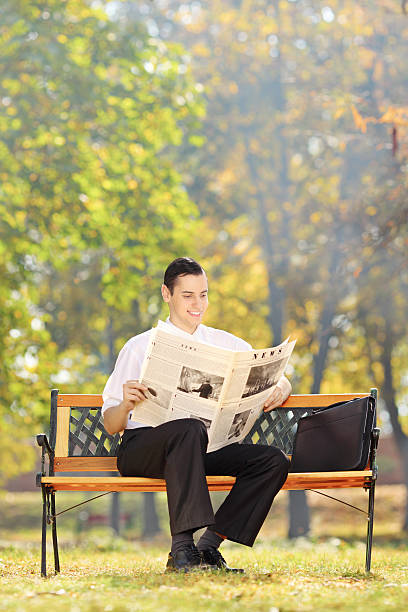 Smiling businessman seated on a wooden bench reading a newspaper in a park