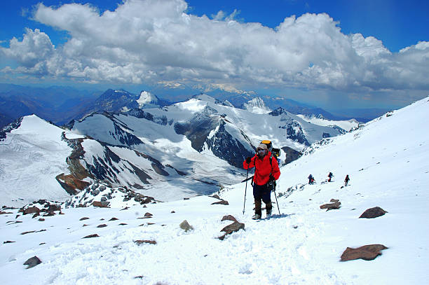 DSLR photo of a group of people hiking on the Perito Moreno glacier. The group is walking on blue ice and are wearing crampons not to slip. They are walking towards the mountain in the background of the picture. It is mostly a blue sky with few clouds. 