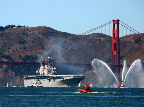 A US aircraft carrier crosses under the Golden Gate Bridge during Fleet Week, led by the Sab Francisco fire boat.  The fireboat shoots streams of water high in the air as a salute to the carrier.  A tug boat is alongside the carrier and a small US Coast Guard skiff patrols nearby.  Hundreds of sailors in white uniforms line the flight deck.