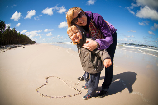Mother with baby in sling and little boy are drawing heart in Baltic sea beach sand.