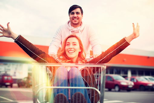 Young Man drive his young woman in a shopping cart outdoors in front of an supermarket. Selective focus to her smiling and outstretching arms.