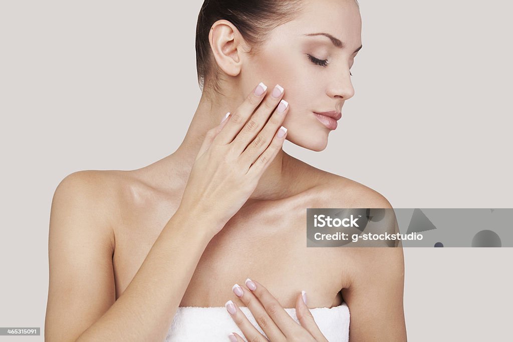 A woman in a towel touching her face Beautiful young woman wrapped in towel keeping eyes closed and touching her face while standing against grey background Moisturizer Stock Photo