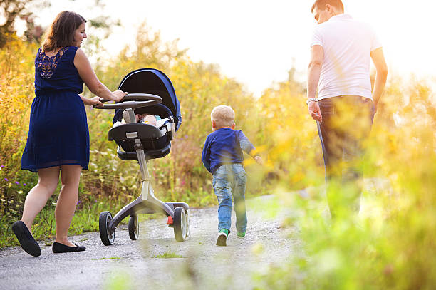 A 3-year-old Japanese girl with twin-tails walks with her 6-month-old baby brother in a stroller and their 30s mother. Captured in early March, in a peaceful rural setting under soft spring sunlight.