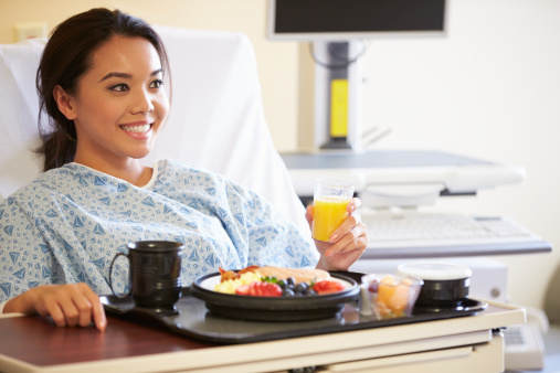 Female Patient Enjoying Meal In Hospital Bed Holding Orange Juice